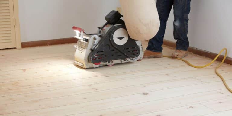 A ReCoat Revolution technician from the knees down using a large sander to remove previous finishes from a hardwood floor before refinishing.