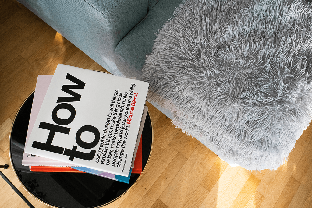 The view of a hardwood floor in a living room from the vantage point of looking down at the corner of a couch and side table with a "How to..." book sitting on top.