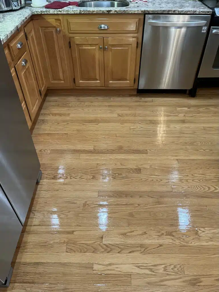 A restored, glossy, stain-free hardwood floor in a residential kitchen after refinishing by ReCoat Revolution.