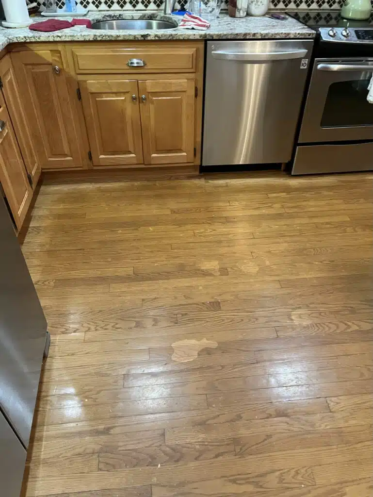 A dull, stained, and scuffed hardwood floor in a residential kitchen before being refinished by ReCoat Revolution.