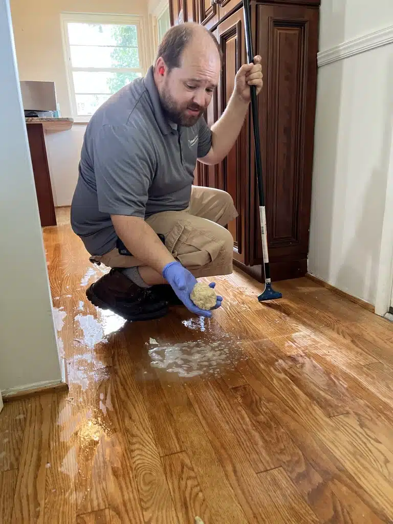 A ReCoat Revolution technician removing the wax buildup from a residential floor, crouched down and holding a palm-sized ball of all of the removed buildup in his gloved hand.