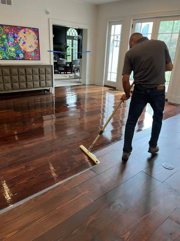 A ReCoat Revolution employee applying new, glossy finish with a foam applicator to a dark, hardwood floor, to protect against damage and provide longevity.