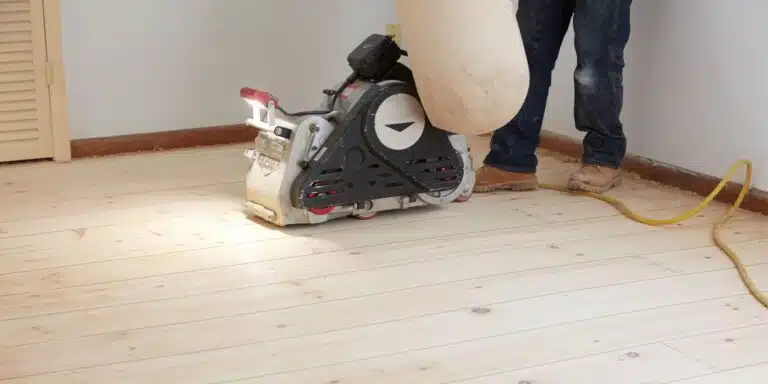 A ReCoat Revolution technician from the knees down using a large sander to remove previous finishes from a hardwood floor before refinishing.