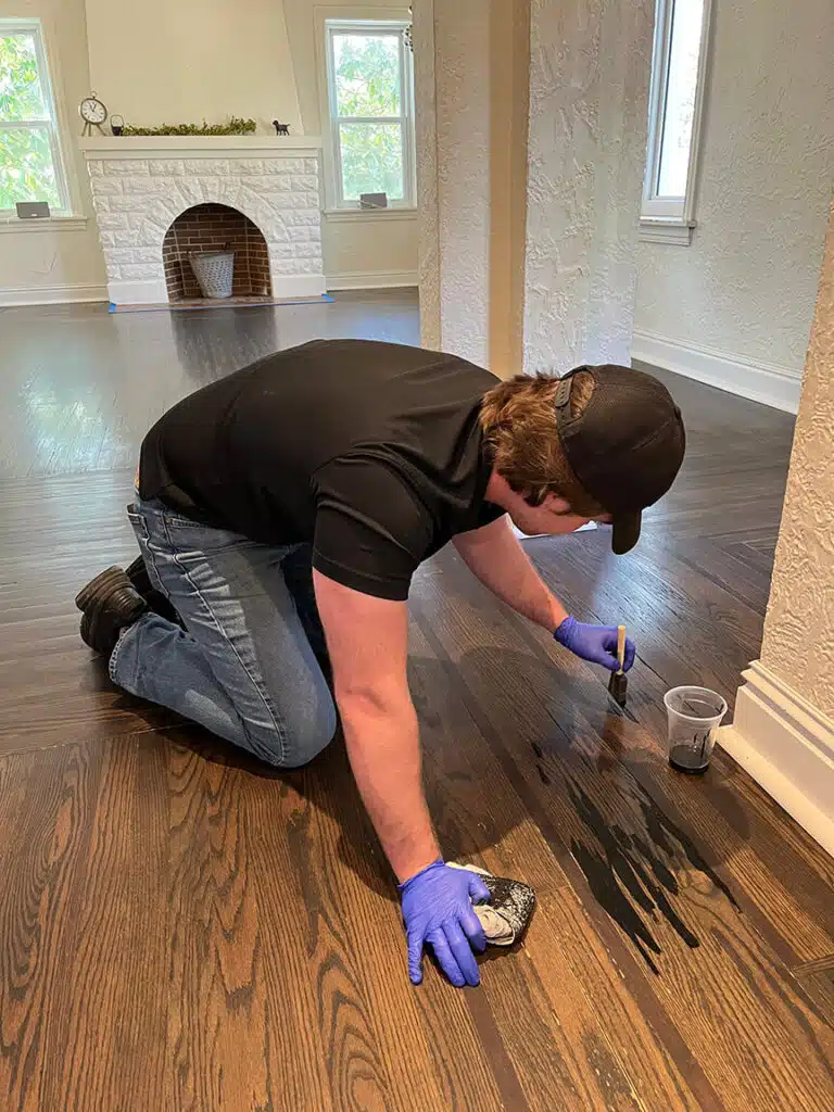 A ReCoat Revolution employee using a small foam brush to apply new, dark stain into floor grooves of the existing hardwood for an even appearance prior to protective coating application.