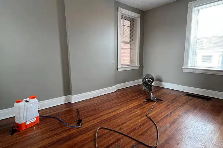 A room within a rental unit that has hardwood floors that is being prepared for refinishing by ReCoat Revolution as a floor sander and solution sprayer sit within the room.