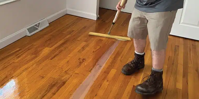 A ReCoat Revolution employee, shown from the waist down, using a sponge tool to spread the new finish on a refinished hardwood floor.
