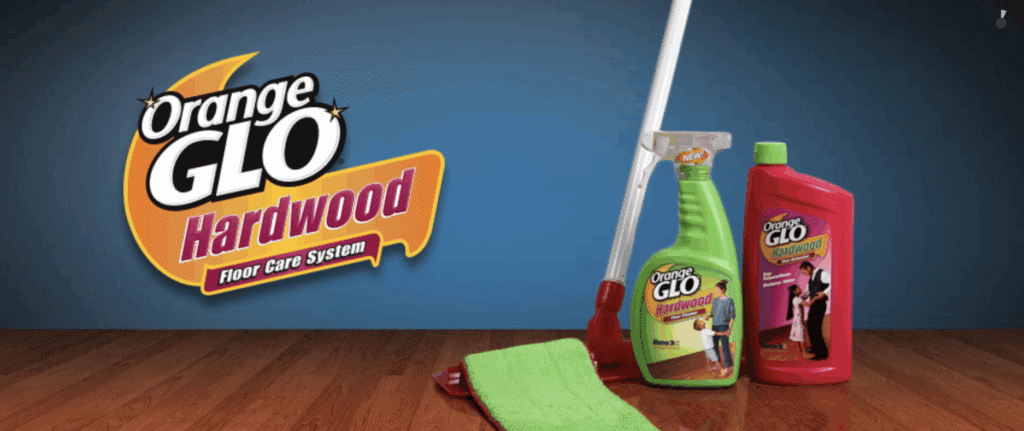 An image displayed as an advertisement for Orange Glo Hardwood Floor Care System, with a green spray bottle product and red product bottle, both sitting near a silver-handled floor cleaning tool with green microfiber cloth for application.