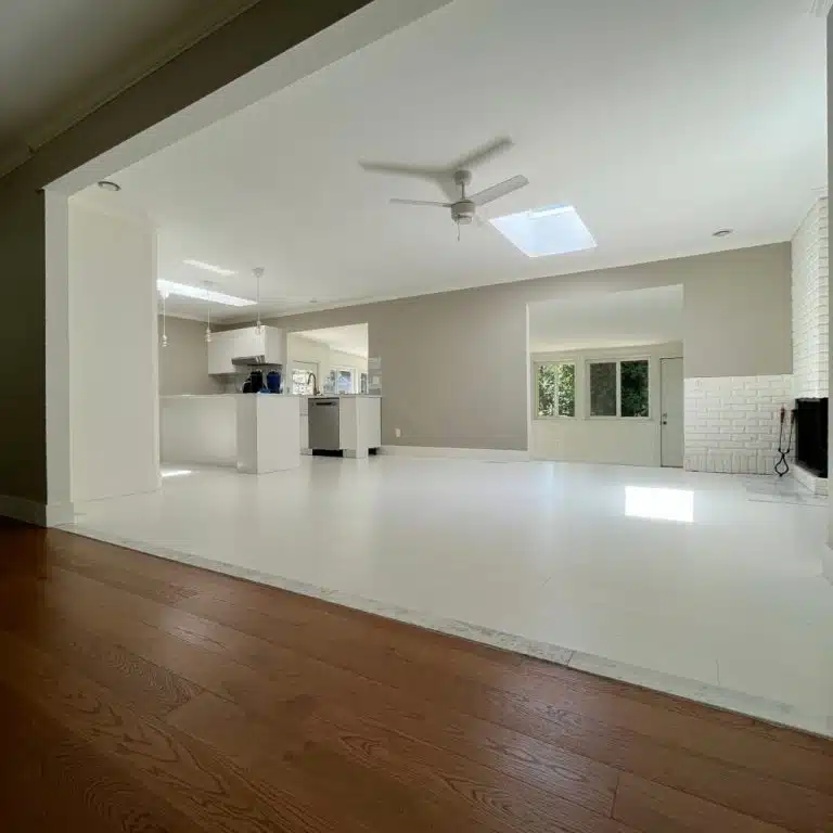 Looking into a bright, all-white kitchen with refinished white flooring by ReCoat Revolution, transitioning out of the room with a stone or marble transition, into natural hardwood.