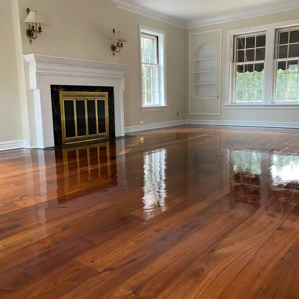 A glossy, refinished hardwood floor completed by ReCoat Revolution in an older home, facing toward a white, built-in fireplace and corner with built-in bookshelf, surrounded by three windows for natural light.