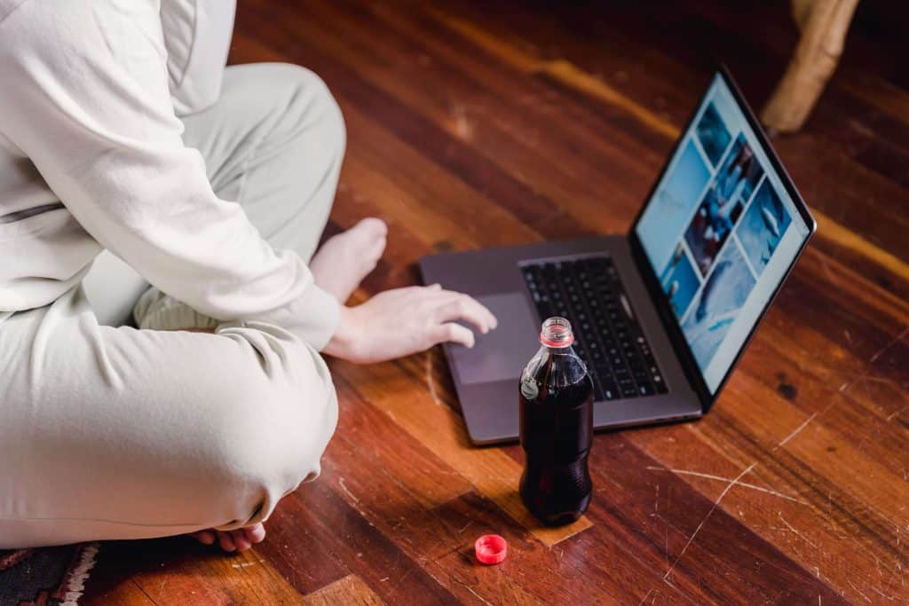 A person sitting cross-legged on a heavily scratched wood floor, scrolling images on a laptop, with an open bottle of dark soda sitting next to the computer.
