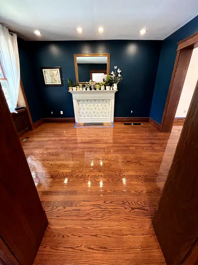 Shining, freshly refinished hardwood floors done by ReCoat Revolution in a home with dark blue walls, a white fireplace, and natural wood trim throughout.