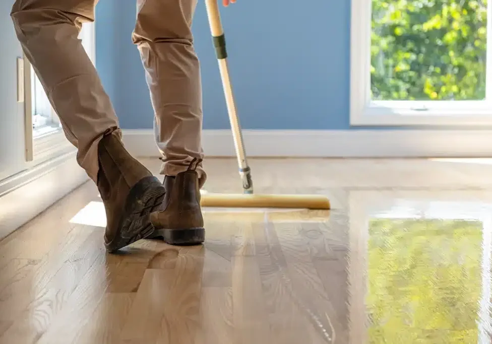 Zoomed in image of a ReCoat Revolution employee's legs from knee down using a foam applicator to spread new finish on a light, hardwood floor.