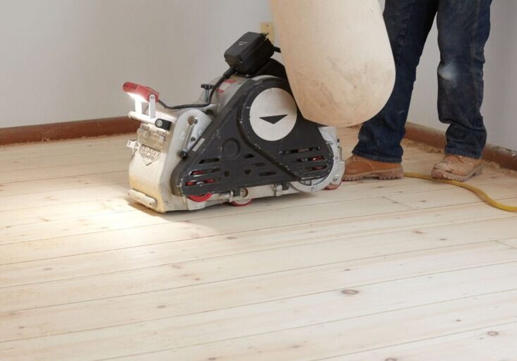 A ReCoat Revolution technician from the knees down using a large sander to remove previous finishes from a hardwood floor before refinishing.