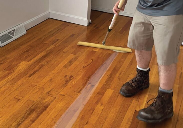A ReCoat Revolution employee, shown from the waist down, using a sponge tool to spread the new finish on a refinished hardwood floor.
